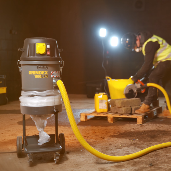 A gray and black Grindex 1100PS wet vacuum cleaner on a wheeled cart, connected by a yellow hose to a yellow bucket in a construction site, with a worker in high-visibility clothing operating a circular saw in the background.